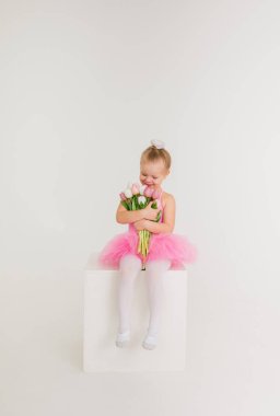 Pembe etekli küçük kız elinde bir buket lale çiçeğiyle beyaz arka planda mesajlaşmak için boşluk bırakarak oturuyor.
