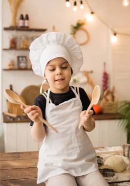 Beyaz önlüklü ve şapkalı neşeli küçük şef mutfaktaki tahta kaşıklarla bir masada oturuyor.