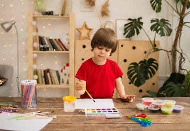 Küçük sanatçı beyaz bir kağıt üzerine fırçayla odadaki ahşap bir masada resim çizer.