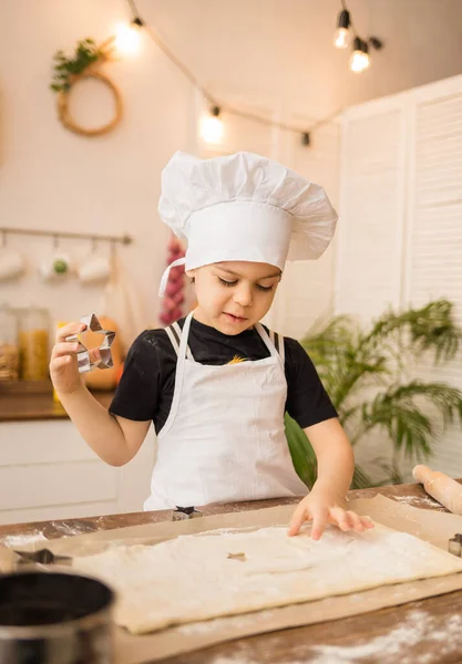 young chef in a white apron and cap cuts cookies out of dough in the kitchen