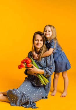 Bir buket kırmızı lale taşıyan mavi elbiseli bir anne sarı arka planda, mesaj atabileceği bir yerde kızının yanında oturuyor. Uluslararası Kadınlar Günü