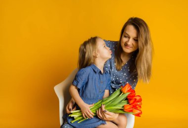 Kırmızı laleli mavi elbiseli kız, sarı arka planda annesini öpüyor. Anneler Günü