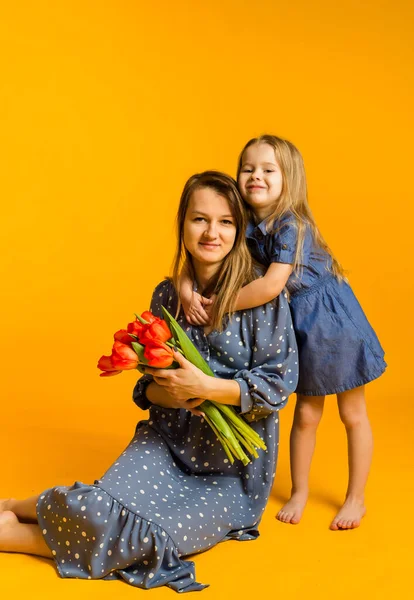 Bir buket kırmızı lale taşıyan mavi elbiseli bir anne sarı arka planda, mesaj atabileceği bir yerde kızının yanında oturuyor. Uluslararası Kadınlar Günü