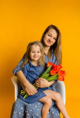 Anne ve kızı bir buket kırmızı laleyle bir sandalyeye oturur ve sarı arka plandaki mesaj için bir yer olan kameraya bakarlar.