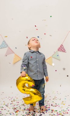 Gömleği ve pantolonu olan küçük bir çocuk iki numaralı altın folyoyu tutuyor ve beyaz arka planda uçan konfetiye bakıyor.