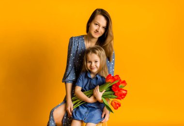 Anne ve kızı bir buket kırmızı laleyle oturur ve sarı arka plandaki kameraya bakar.