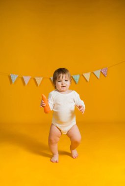 Beyaz elbiseli küçük bir kız sarı arka planda havuç ve çelenkle yürüyor.