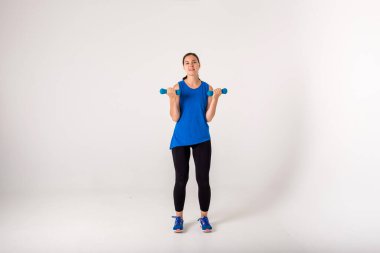 Kadın halterlerle egzersiz yapıyor ve beyaz arka plandaki kameraya bakıyor.