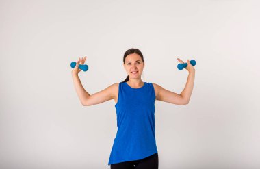 Dambıllarla el egzersizi yapan ve beyaz arka plandaki kameraya bakan bir kadının portresi..