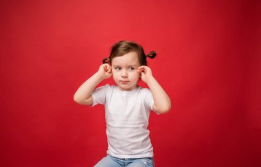 Genç kız kulaklarını parmaklarıyla kapadı. Kırmızı arka planda beyaz tişörtlü bir kız.