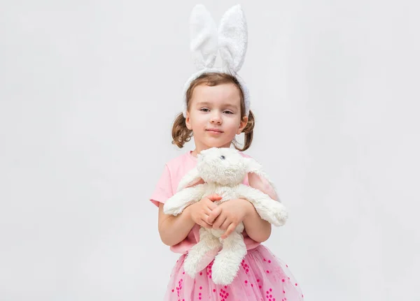 Beyaz arka planda, boş bir koltuğu olan tatlı bir kız. Tavşan kulaklı ve oyuncak tavşanlı bir kız. Pembe tişörtlü küçük bir kız. Kız kameraya bakıyor.