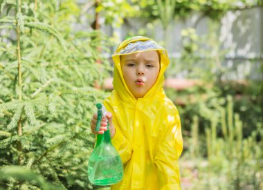 Sarı yağmurluk giymiş, elinde sprey silahla kozalaklı bir serada küçük bir kız kameraya şaşkın bir ifadeyle bakıyor.