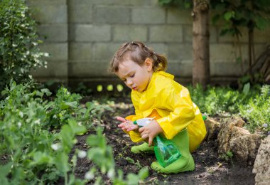 Sarı yağmurluk ve yeşil lastik çizmeli küçük bir kız asistan bahçedeki bitkilerin sulanmasına yardım ediyor.
