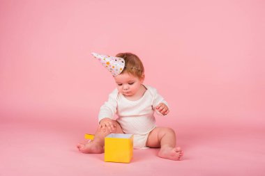 Şenlik şapkalı küçük kız pembe arka planda sarı bir hediye kutusu açıyor.