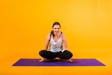 Sporcu kadın, Tolasan pozunda mor bir Mat 'ın üzerinde oturuyor. Turuncu bir arka planda, uzayın bir kopyasıyla.