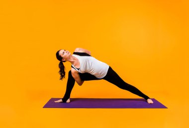 Yogacı kadın turuncu arka planda mor bir Mat üzerinde poz veriyor.