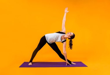 Bir kadın turuncu arka planda mor bir Mat üzerinde Parivritta Trikonasana performansı sergiliyor.