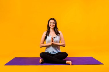 Bir kadın elleri Namaste 'de katlanmış, turuncu bir arka planda mor bir mindere oturur.