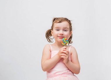 Pembe elbiseli, beyaz bir arka planı olan, at kuyruklu tatlı bir kız. Lolipopu olan sarışın bir kız. Kız ileriye doğru gülümsüyor.