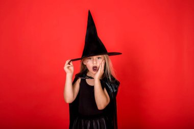 Cadılar Bayramı 'nda kırmızı izole edilmiş bir arka planda cadılar bayramı şapkalı bir cadının görüntüsüne şaşırmış bir kız.