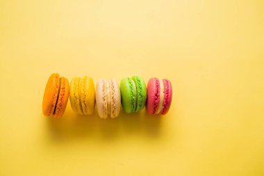 Sarı arka planda bir dizi makaron. Makaronlar sarı, turuncu, krema, yeşil ve pembedir. Üst üste beş tane badem kurabiyesi istiflenmiş. opi alanı.