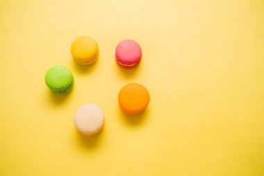 Sarı arka planda bir dizi makaron. Makaronlar sarı, turuncu, krema, yeşil ve pembedir. Beş badem kurabiyesi bir çemberde toplanmış. ? opy boşluk.