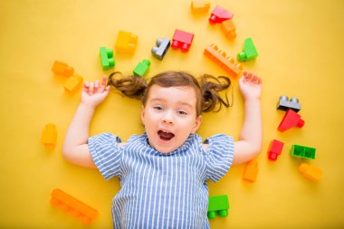 Küçük bir kız inşaat setiyle sırt üstü yatıyor. Sarı arka planda sürpriz bir kız. Soyun. Çizgili gömlekli bir kız kameraya bakıyor.