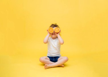 Tatlı bir kız oturuyor ve portakal parçalarını gözlerinin önünde tutuyor. Küçük bir kız bacak bacak üstüne atmış oturuyor. Sarı arka planda beyaz tişörtlü ve kot şortlu küçük bir kız..