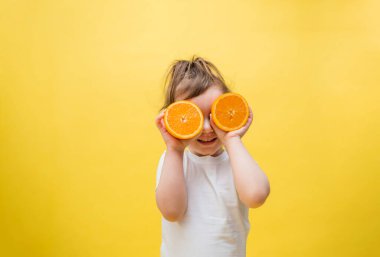 Tatlı bir kız portakalların yarısını gözlerinin önünde tutuyor. Sarı arka planda beyaz tişörtlü küçük bir kız..