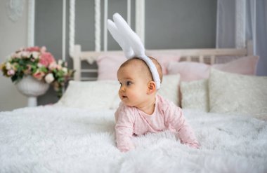 Saç bandı takmış tavşan kulaklı bir kız bebek odadaki beyaz bir battaniyenin üzerinde sürünüyor.