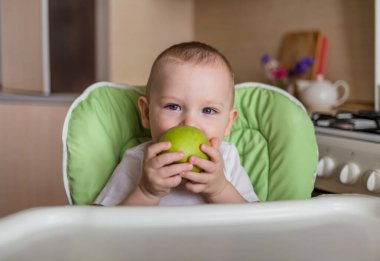 Bebek bir sandalyeye oturup yeşil elma yiyor.