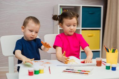 Küçük bir kız ve bir oğlan bir masaya oturur ve avuçlarını kağıda çizerler.