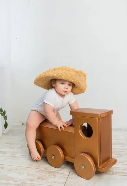 little girl in a white bodysuit and a straw hat sits on a wooden locomotive on a white background