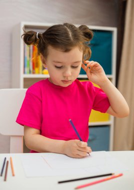 Düşünceli küçük kız odadaki masada renkli bir kalemle kağıt üzerine resim çiziyor.