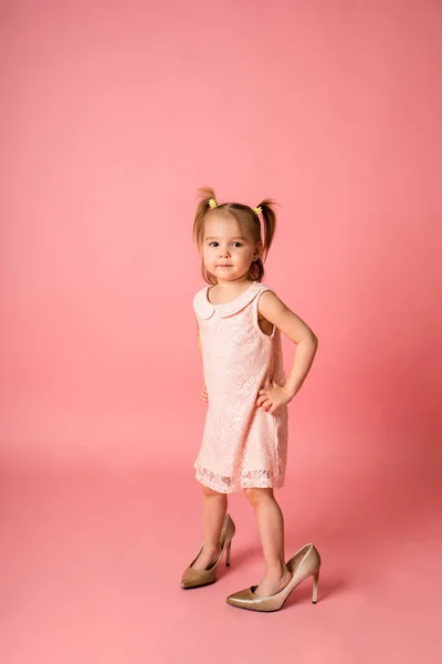 Pembe dantel elbiseli küçük bir kız ve annesinin ayakkabıları pembe bir arka planda duruyor.