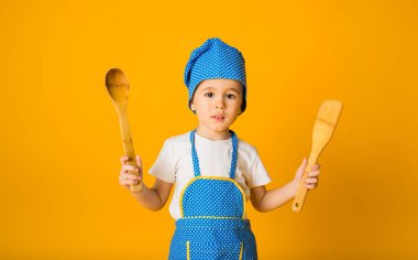 Sarı arka planda ahşap kaşıklarla ahşap şapkalı ve önlüklü küçük bir çocuğun portresi.