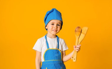 Şef kostümü giymiş küçük bir çocuk sarı arka planda ahşap kaşıklar tutuyor ve mesaj için yeri var.