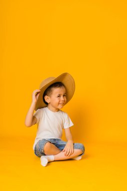 Hasır şapkalı neşeli çocuk sarı arka planda oturuyor ve başka tarafa bakıyor.