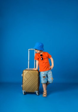 Tişörtlü, şortlu ve panama şapkalı küçük bir çocuk mavi bir arka planda mesaj için yeri olan bir bavul tutuyor.