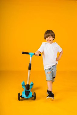Beyaz tişörtlü ve kot şortlu küçük bir çocuk sarı arka planda scooter tutuyor ve mesaj için yeri var. Çocuklar için spor