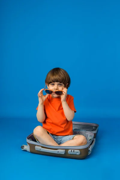 Kahverengi saçlı küçük beyaz çocuk güneş gözlükleriyle oynuyor ve mavi arka planda bir bavulda oturuyor.