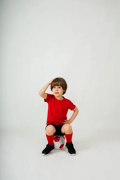 Задумчивый Маленький Мальчик Спортивной Форме Сидит Футбольном Мяче Белом Фоне — стоковое фото