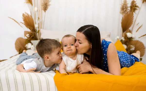 ブルネットの母親と息子は部屋のベッドで彼らの小さな娘にキスをする 幸せな子供時代 — ストック写真