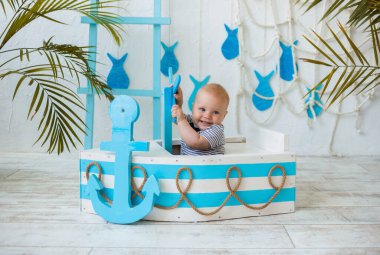 Çizgili tulumlu mutlu bebek tahta bir teknede oturur ve direksiyonu deniz dibinde tutar. Yaz seyahati