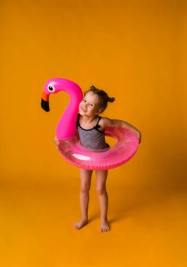 Çizgili mayo giymiş neşeli küçük bir kız flamingo şeklinde şişme bir yüzme çemberi olan sarı bir arka planda duruyor ve mesaj için bir yer var. Yaz tatili