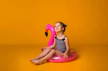 Çizgili mayo giymiş küçük bir kız flamingo ile şişirilebilir pembe bir çemberin üzerinde oturuyor ve sarı bir arka planda mesaj için yeri olan bir deniz tatili hayali kuruyor. Yaz çocuğu