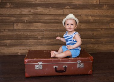Çizgili tişörtlü ve hasır şapkalı küçük bir çocuk ahşap bir arka planda, eski bir bavula oturmuş, mesaj için yeri var. Yaz seyahati