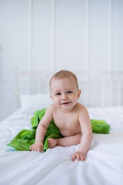 Güzel erkek bebek yeşil havluyla beyaz bir yatakta oturur ve kameraya bakar. Dikey yönelim