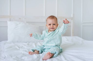 Çizgili pijamalı sevimli küçük çocuk beyaz bir yatakta oturur ve kameraya bakar. Sağlıklı bebek uykusu