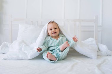 Çizgili pijamalı neşeli küçük bir çocuk odada beyaz bir yatakta oturuyor ve oynuyor. Mutlu çocukluk